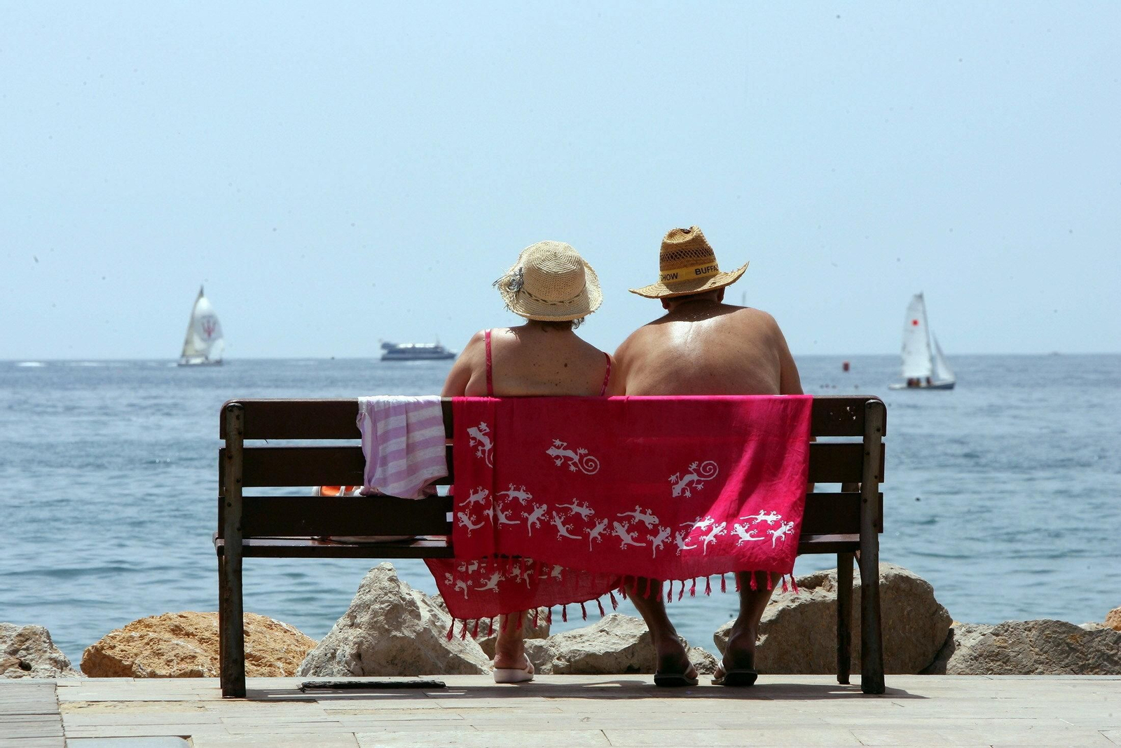 Una pareja de jubilados contempla el mar mientras toma el sol en un banco.
