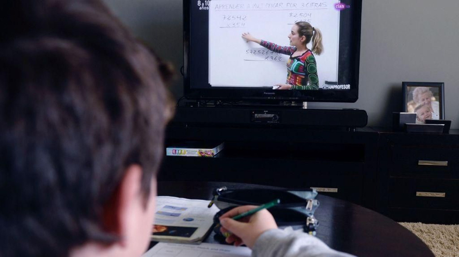 Un niño sigue desde su casa las indicaciones de una profesora a través de la pantalla del televisor.