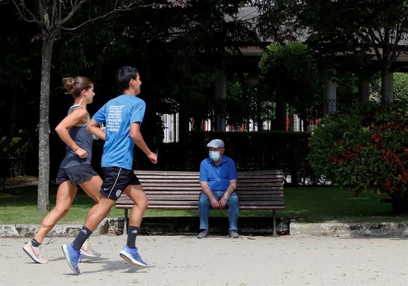 Un hombre con mascarilla descansa en un banco mientras una pareja de corredores practica deporte en el Parque de Rosalía de Castro en Lugo