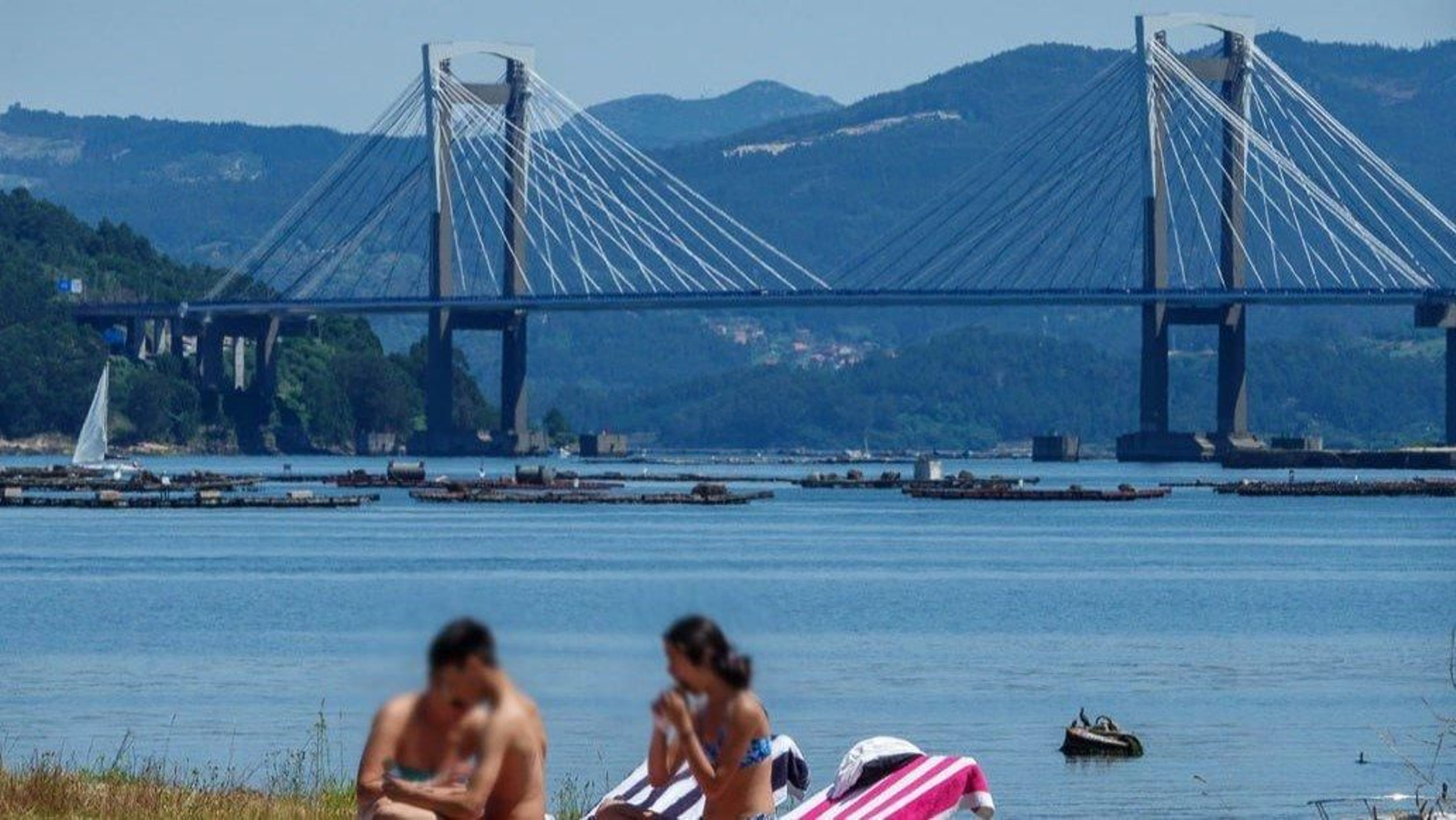 Unos jóvenes disfrutan del buen tiempo con el puente de Rande al fondo. // Vicente Alonso