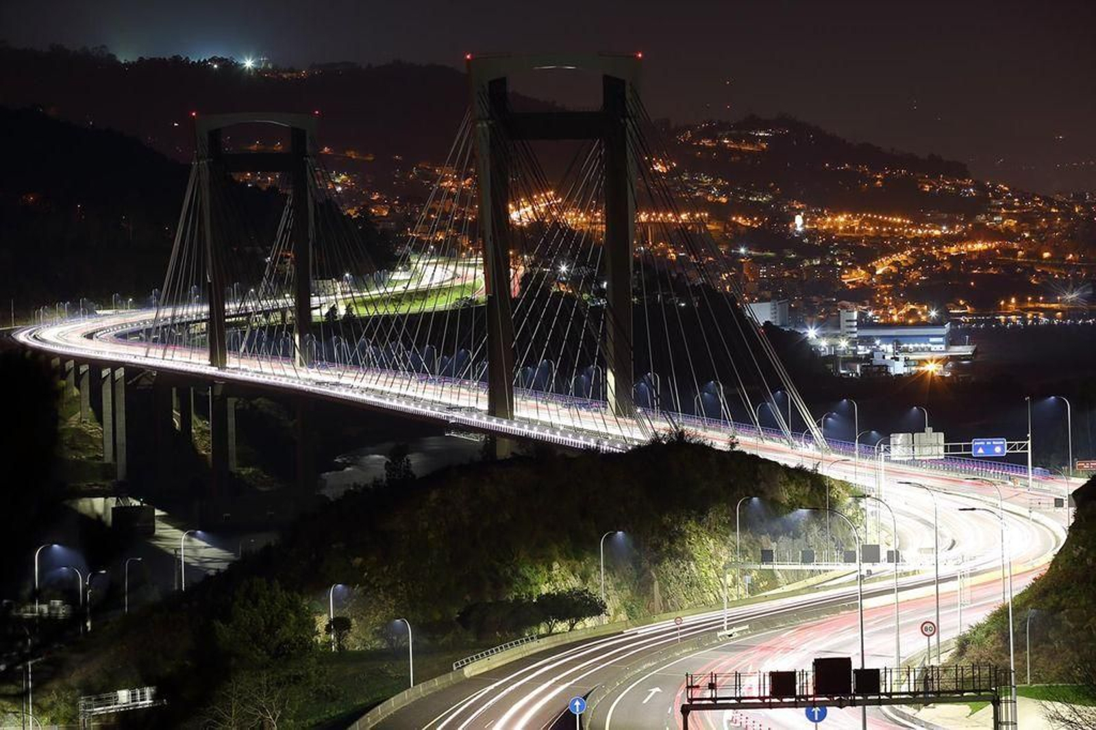 El puente de Rande tendrá una iluminación ornamental y de momento ya se puede observar su perfil de noche mediante un truco fotográfico en una imagen tomada desde O Morrazo, con la ciudad de Vigo al fondo, al revés de lo habitual.
