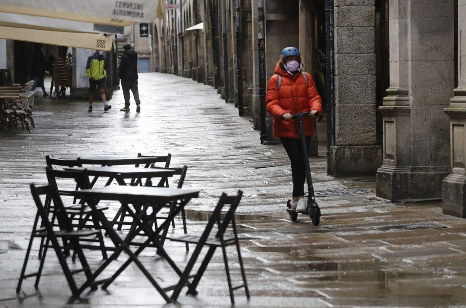 Una joven circula en patinete ante una terraza vacía de una cafetería de Santiago.