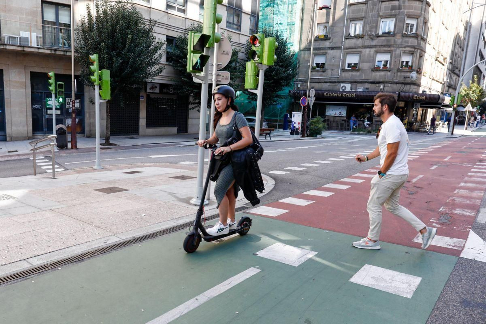 Una usuaria de un patinete eléctrico en el carril bici junto a un peatón cruzando la calle.