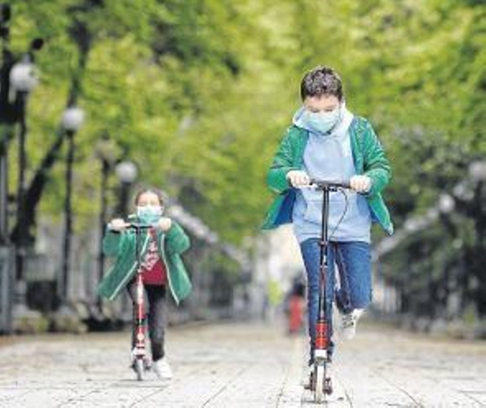 Dos niños juegan que sus patinetes en un parque, protegidos con mascarillas.