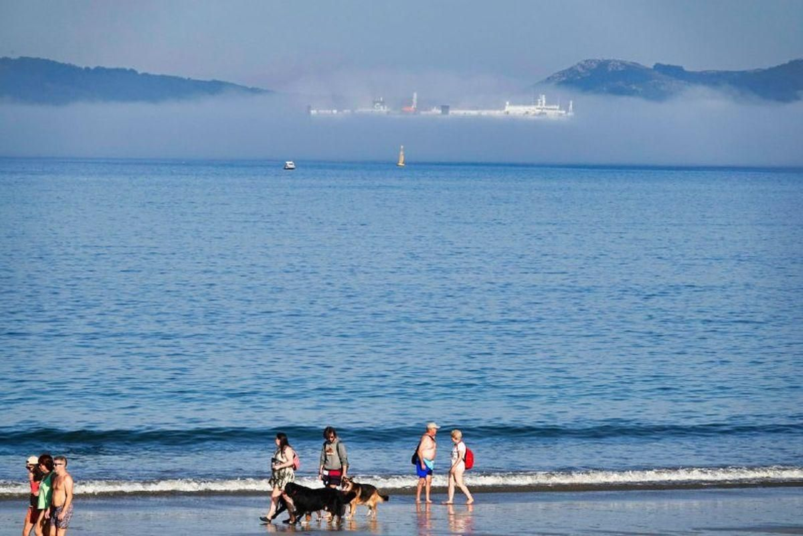 Bañistas paseando por la playa, con las Cíes al fondo, cubiertas por un manto de niebla.