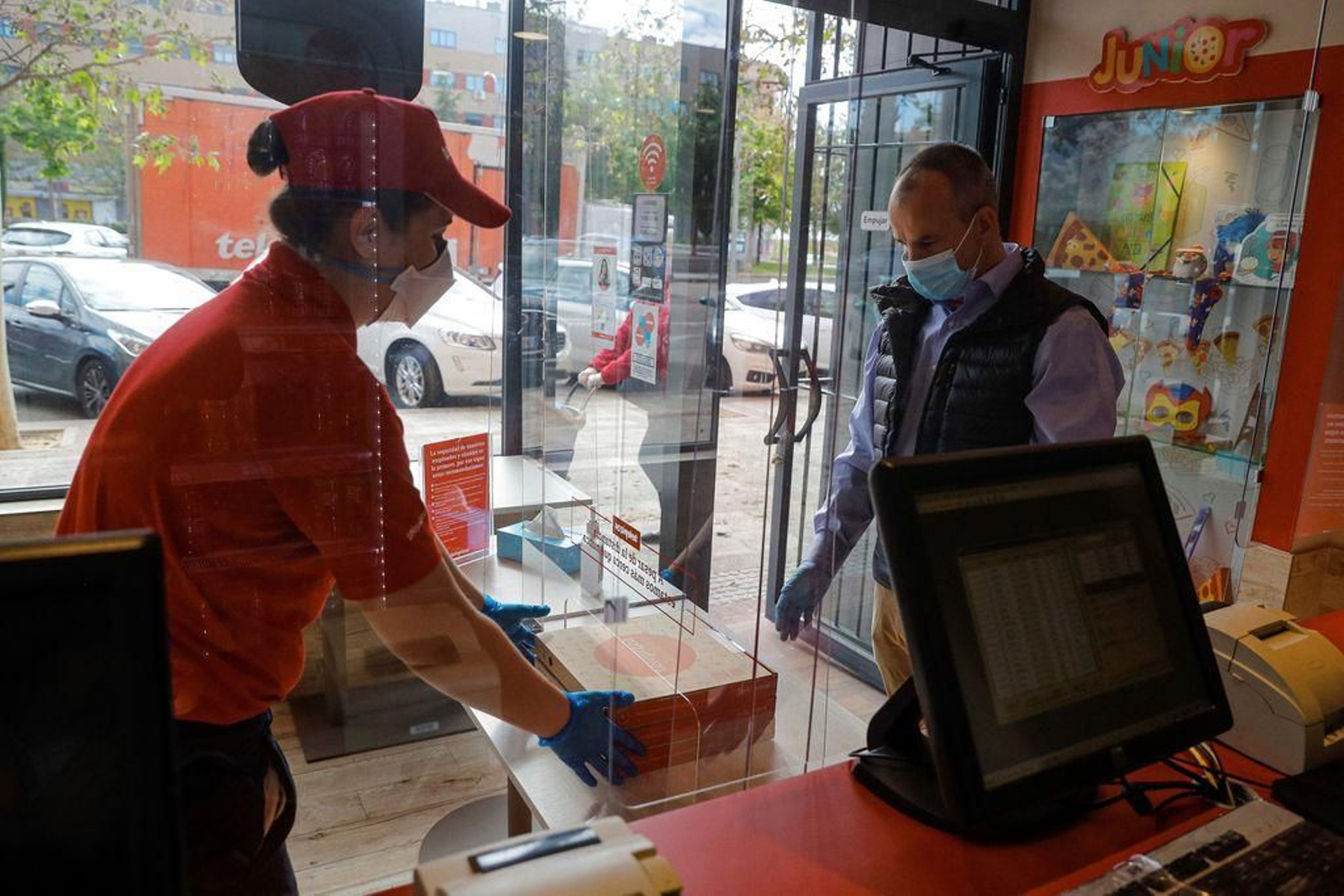 Una empleada de una pizzería entrega un pedido a un cliente tras una mampara.