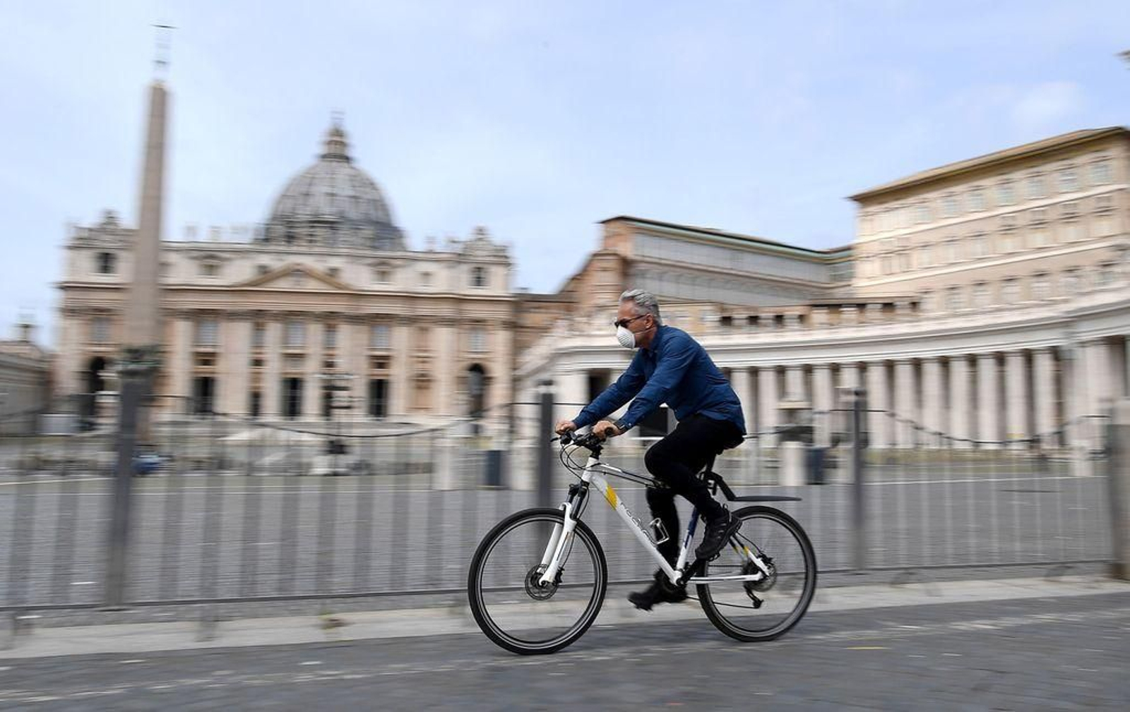 Un hombre con mascarilla viaja en bicicleta por Roma, con el Vaticano de fondo.
